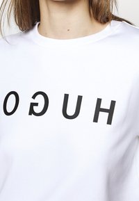 Vit bomullströja med en svart omvänd "HUGO"-logotyp tryckt över framsidan. Rund halsringning och mjuk textur.