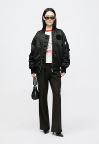 Crna bomber jakna s patentnim zatvaračem, okrugli pečat, uparena s bijelom majicom s grafikom, smeškim hlačama širokih nogavica i crnim dodacima.