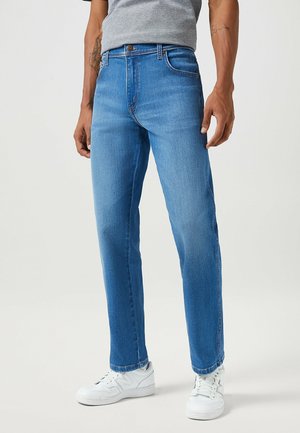 Homme portant un jean droit bleu clair, un t-shirt gris et des baskets blanches, debout devant un fond uni.
