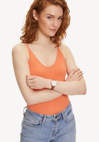Oranges, geripptes Bodysuit mit dünnen Trägern, kombiniert mit hellblauen, hochgeschnittenen Jeans und einer braunen Uhr mit Lederarmband. Einfarbiger Hintergrund.