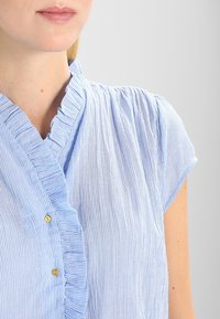 Light blue, striped blouse with a ruffled collar and short sleeves. Gold button detail at the neckline. Textured fabric with vertical lines.
