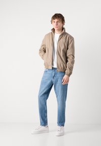 Lys beige bomberjakke med høj krave, elastiske ærmer og talje, parret med lys blå jeans og hvide sneakers.
