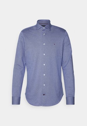 Chemise à manches longues boutonnée en tissu bleu texturé avec un col pointu et un petit logo brodé sur la poitrine.
