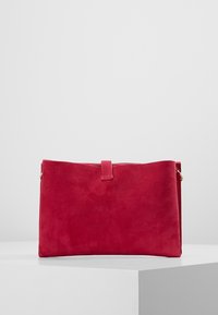 Punainen mokkapintainen käsilaukku, jossa on sileä pinta, päällä vetoketju ja pieni lenkki helppoa käsittelyä varten. Yksinkertainen, koristamaton muotoilu.