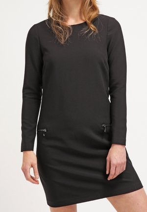 Femme portant une robe noire à manches longues, longueur genoux, avec deux poches zippées horizontales près des hanches.