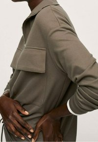 Sudadera color caqui con cuello con cremallera, dos grandes bolsillos delanteros y detalles blancos en los puños. Confeccionada en tela suave y texturizada.