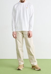 Man in een wit overhemd met lange mouwen, beige broeken en beige sneakers, staande op een groene vloer tegen een effen witte muur.