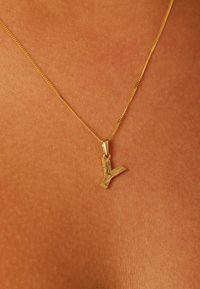 Collier en or avec un pendentif en forme de Y, orné de gravures détaillées. La chaîne est délicate et lisse, posée sur un fond de peau texturée.