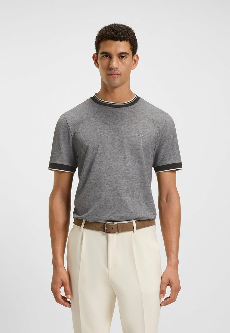 T-shirt grigio a maniche corte con colletto e dettagli delle maniche in contrasto bianco e nero, indossato con pantaloni color crema e una cintura marrone.