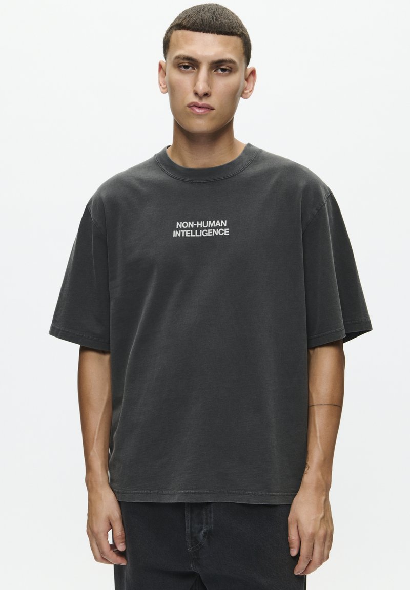 T-shirt noir surdimensionné en mélange de coton avec des manches courtes, affichant un texte blanc "INTELLIGENCE NON HUMAINE" sur la poitrine, à col rond.