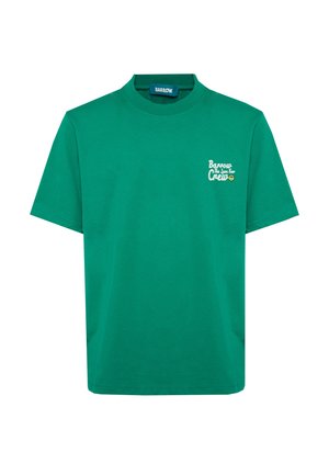 Giovane uomo con capelli ricci che indossa una maglietta verde, pantaloni grigio chiaro, scarpe da ginnastica bianche, in piedi con una mano in tasca e tatuaggi visibili sulle braccia.