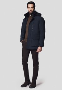 Granatowa puchowa kurtka z odpinanym kapturem, z przodu na zamek i wieloma kieszeniami, noszona na brązowym swetrze i brązowych spodniach.