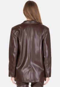 Donna con capelli lunghi che indossa un blazer in pelle marrone scuro e pantaloni abbinati, vista da dietro su uno sfondo semplice.