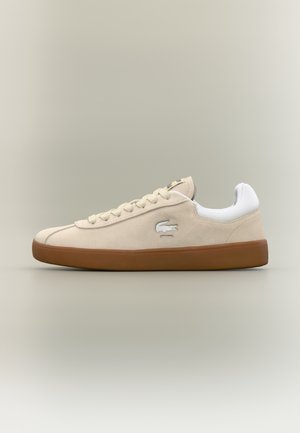 Baskets basses beige avec semelle marron, talon blanc rembourré, lacets à l'avant et logo crocodile sur le côté.