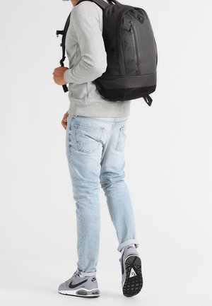 Mand iført lyseblå jeans, grå sweatshirt, grå Nike sneakers og bærer en stor sort rygsæk, går væk på en hvid baggrund.