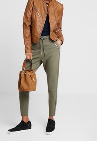 Hnědá kožená bunda se zipovými kapsami, námořnické tričko, olivově zelené tepláky se bočními pruhy, béžová kabelka a černé slip-on tenisky.