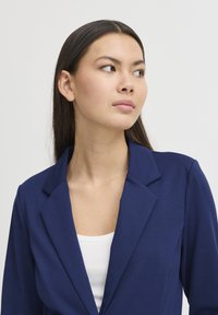 Blazer azul marino con solapa, hecho de una tela suave. Llevado sobre una blusa blanca, mostrando un corte entallado y líneas limpias.