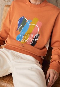 Sudadera naranja con un contorno blanco de un caballo y patrones de pinceladas coloridas. Mangas y dobladillo acanalados, material de algodón. Combinada con pantalones blancos.