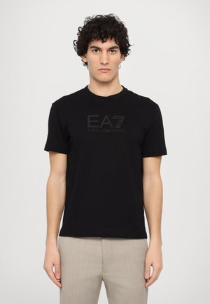 T-shirt in cotone nero con scollatura rotonda, maniche corte e stampa opaca del logo "EA7 Emporio Armani" sul davanti, abbinata a pantaloni beige chiaro.