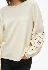 Felpa color crema con maniche lunghe, caratterizzata da motivi floreali tagliati al laser sui polsini. Tessuto morbido e vestibilità comoda.