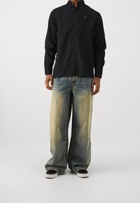 Camisa negra de manga larga con botones y un logo sutil, combinada con jeans de pierna ancha en azul claro y desgastados, finalizada con zapatillas negras.