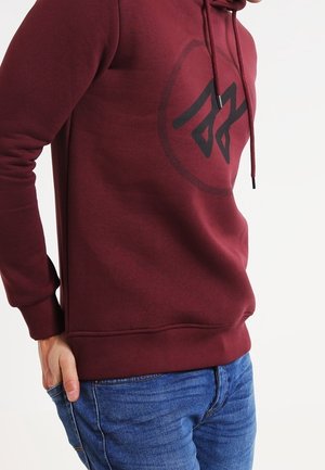 Mies pukeutuneena burgundinpunaiseen huppariin, jossa musta abstrakti logo, ja sinisiin farkkuihin, kädet taskussa yksiväristä valkoista taustaa vasten.