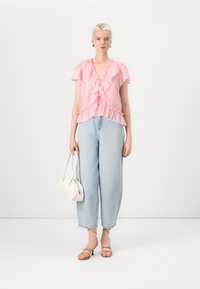 Top rosa con volant e maniche corte, abbinato a jeans azzurri larghi e sandali beige con cinturini. Nella mano tiene una piccola borsa bianca.