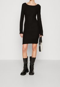 Černé bodycon šaty s dlouhým rukávem a kulatým výstřihem, doplněné černými vysokými kozačkami a malou černobílou kabelkou. Hladký povrch.