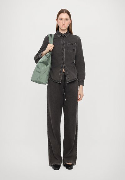 Camisa de ganga preta com fecho de botão e bolso no peito, combinada com calças pretas de corte largo. A segurar uma mochila de cordão verde-claro.