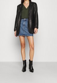 Blazer en cuir noir sur un top vert col en V, associé à une mini-jupe en denim bleu et des bottines à talons noirs. Textures lisses et formes ajustées.