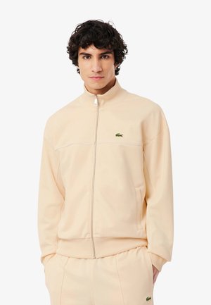 Jonge man met krullend haar, gekleed in een beige jas met rits en bijpassende broek, met het groene krokodillenlogo zichtbaar op beide kledingstukken.