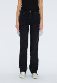 Schwarze Denim-Jeans mit geradem Bein, fünf Taschen, Knopfverschluss und dezenten Steppdetails. Tragen mit hellen Sneakers.
