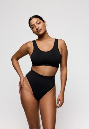 Vrouw met donker haar in een zwarte sportbeha en hoogwaterondergoed die poseert tegen een effen lichte achtergrond.