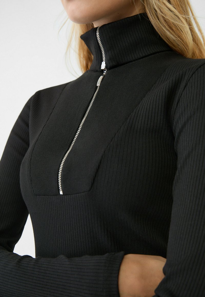 Il top nero a costine con colletto alto presenta una mezza zip con hardware in argento. Il design include un frontale a pannelli e maniche lunghe attillate.