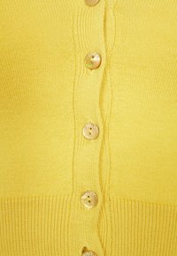 Gul kardigan med ribbat mönster och rund halsringning, fäst med runda, ljusa knappar längs framsidan.