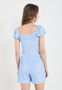 Blå blommig jumpsuit med puffärmar, fyrkantig halsringning och strukturerad tyg. Har ett elastiskt midjeband och shorts.