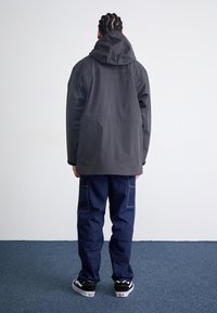 Zwarte waterdichte jas met een capuchon, gecombineerd met donkerblauwe cargobroeken en zwarte sneakers. Glad materiaal, casual ontwerp met een relaxte pasvorm.