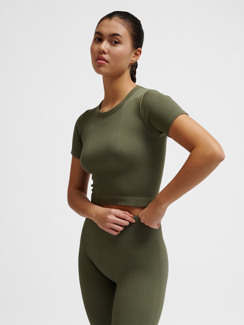 Femme ajustant un ensemble crop top et leggings taille haute côtelé vert olive, debout devant un fond clair uni.