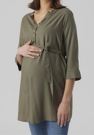 Donna incinta con tunica verde oliva annodata e jeans blu, che tiene delicatamente la pancia su uno sfondo grigio uniforme.