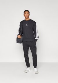 Conjunto de desporto masculino preto com detalhes em branco nas três riscas. Composto por um sweatshirt de gola redonda e calças de fato ajustadas, acompanhado por sapatilhas brancas.