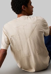 T-shirt in cotone beige con grande logo ricamato "CK" sul retro. Maniche corte e vestibilità casual, con una texture liscia.