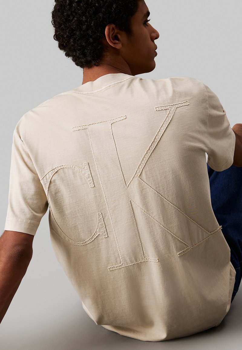 T-shirt en coton beige featuring un grand logo "CK" brodé dans le dos. Manches courtes et coupe décontractée, avec une texture lisse.