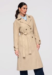 Femme portant un trench-coat beige à double boutonnage avec ceinture, chemise bleu clair, pantalon foncé, boucles d'oreilles dorées et cheveux foncés attachés en queue de cheval basse.