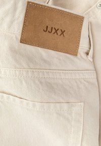 Pantalones de mezclilla beige con parche de cuero cuadrado cosido de la marca etiquetado "JJXX" sobre el bolsillo trasero y presilla para cinturón visible.