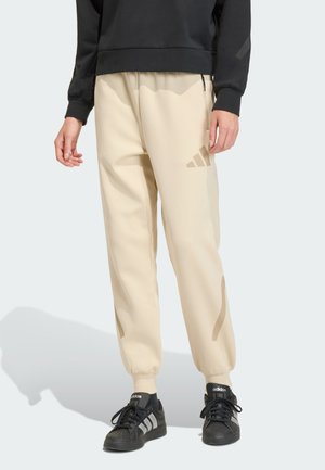 Osoba ubrana w beżowe spodnie dresowe Adidas oraz czarne buty Adidas, stojąca z ramionami swobodnie opuszczonymi wzdłuż ciała na białym tle.