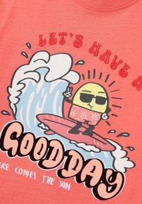 Camiseta color coral con un gráfico que presenta un personaje de dibujos animados surfeando una ola, con gafas de sol y el texto que dice "TENGAMOS UN BUEN DÍA".