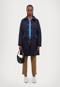 Jūras spēku piegrieztā garā mīkstā mētelī ar pogām, sānu kabatām, apvienots ar gaiši zilu džemperi, brūnām biksēm, baltām sporta apaviem un melnu rokassomu.