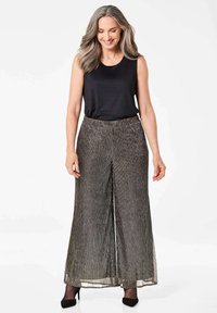 Zwarte mouwloze top gecombineerd met wijdvallende, geplooide metallic zilveren broek. De stof heeft een gestructureerde glans. Model draagt zwarte hakken.