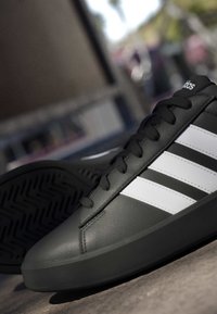 Baskets Adidas noires avec trois bandes blanches et lacets noirs, montrées sous un angle avec un arrière-plan urbain flou.