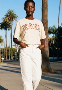 Naine beežis lühikeses T-särgis, millel on tekst "KANU", ja kõrge vöökohaga valgetes pükstes seisab õues taustal palmipuud ja selge taevas.